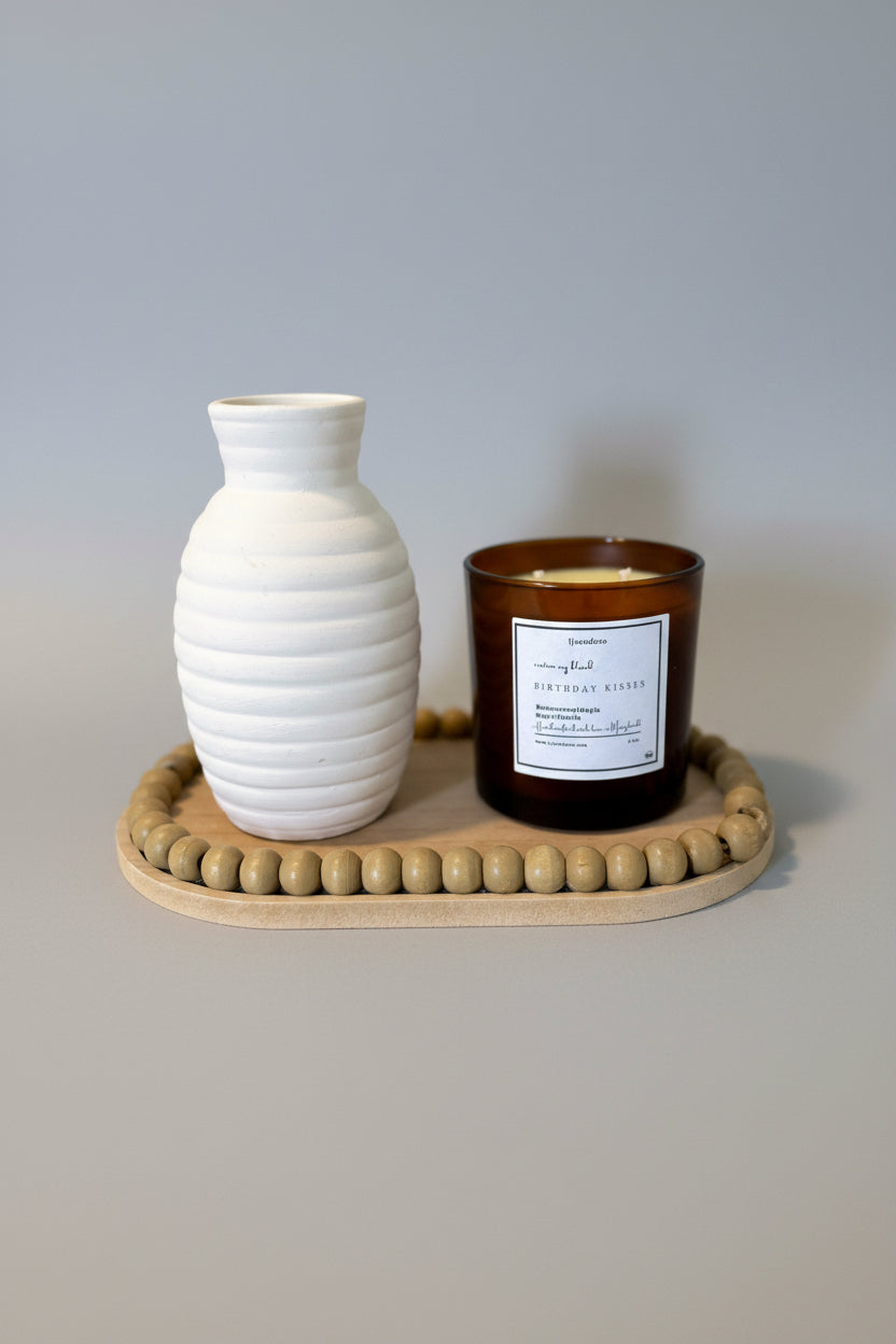 A white ceramic vase and a brown wooden candle holder with a scented candle on top, placed on a wooden surface with a beige wall in the background.