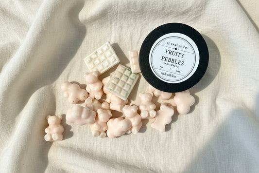 Candle labeled 'Fruity Pebbles' surrounded by small, pastel-colored candy-like objects on a light fabric background.